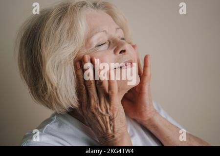 Portrait of smiling beautiful senior woman touching her cheek, after using anti-aging cosmetics in the morning isolated over gray background. People Stock Photo