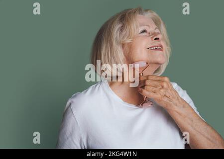 Lovely senior woman doing jade roller facial massage stimulating lymphatic drainage, isolated over green background. Spa technique skincare ads. Anti Stock Photo