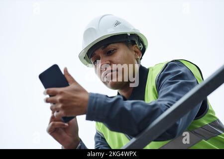 Low angle portrait of female worker wearing hardhat and using smartphone against sky Stock Photo