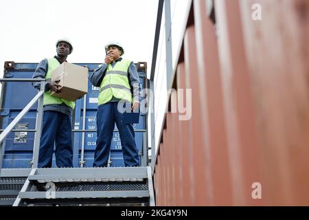 Full length portrait of two workers standing on container at shipping docks, copy space Stock Photo