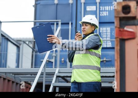 Portrait of female foreman wearing hardhat and speaking to radio while managing operations at shipping dock Stock Photo