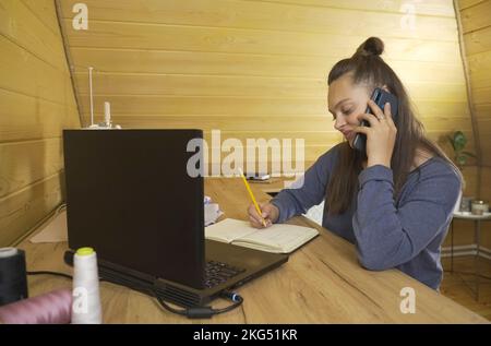 Woman fashion designer working on a laptop, making notes in a notebook, talking on the phone with a client. Small business. Stock Photo
