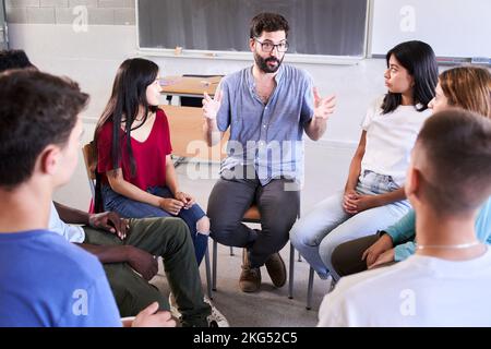 Diverse group of high school students sitting on chairs in a circle and interacting during a lesson, their Caucasian male teacher with them and Stock Photo