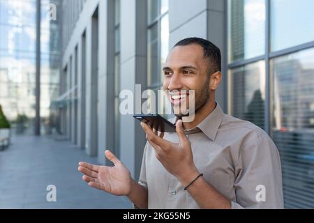 Young handsome man hispanic, african american standing outside, holding phone, talking through loudspeaker, recording on phone, smiling. Stock Photo