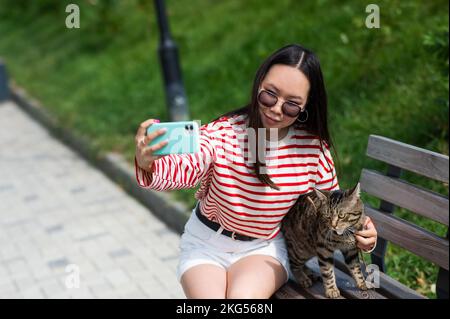 Young woman sits on a bench with a tabby cat and takes a selfie on a smartphone outdoors.  Stock Photo