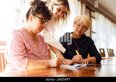 Young smiling woman businesswoman standing behind two elderly women sitting at desk, explaining business plan in office. Stock Photo