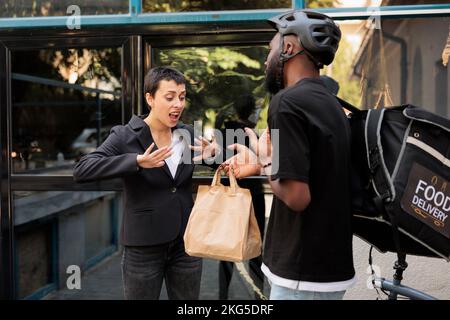 Office food delivery delay, impulsive customer getting wrong order. African american deliveryman giving angry unsatisfied customer takeaway meal in front of company building outdoors Stock Photo