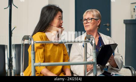 Physician using digital tablet to consult patient with cervical neck brace, sitting in waiting area lobby. Attending medical appointment with doctor, recovering after fracture accident. Stock Photo