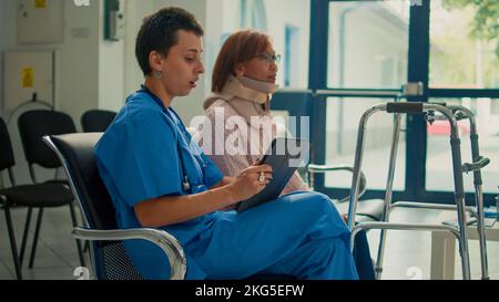 Nurse consulting patient with neck collar brace and walking frame, taking notes about accident injury on tablet. Doing checkup visit with woman wearing cervical foam and impairment. Stock Photo
