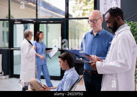 Physician medic showing illness diagnosis report to senior patient discussing health care treatment during examination in hospital waiting room. Old man fillling medical papers, medicine concept Stock Photo