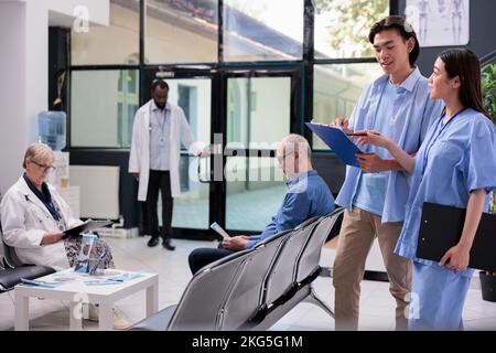 Asian young patient holding clipboard filling medical report before start consultation with medic during checkup visit in hospital waiting area. Diverse people standing in reception, medicine service Stock Photo