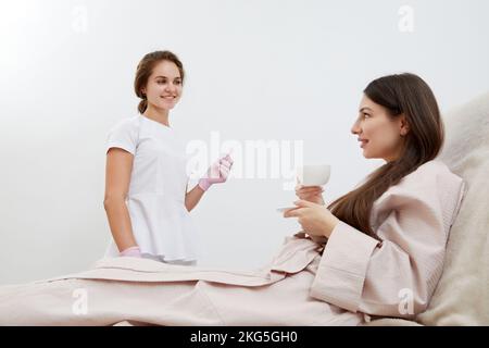 Beautician serving cup of coffee to her client after body treatment in beauty salon. Relaxing with beverages on a cosmetic bed. Beauty , pleasure Stock Photo