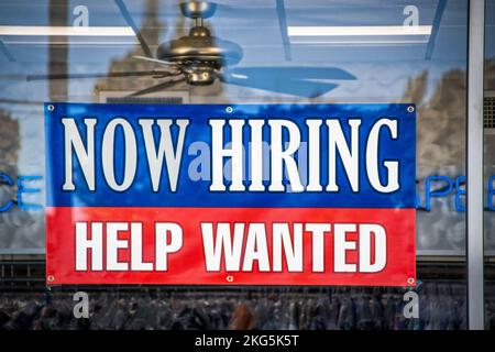 Now Hiring Help Wanted Sign in window of business with ceiling fan visible inside Stock Photo