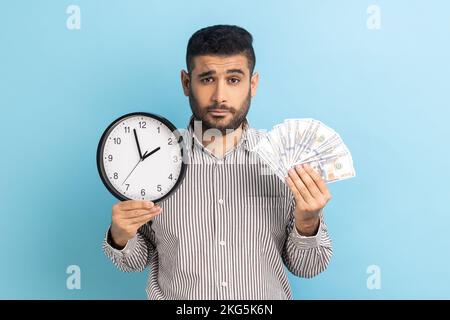 Portrait of upset sad businessman holding big fan of dollar banknotes and wall clock, time is money, looking at camera, wearing striped shirt. Indoor studio shot isolated on blue background. Stock Photo
