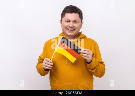 Satisfied positive middle aged man holding German flag in her hand and looking at camera with toothy smile, wearing urban style hoodie. Indoor studio shot isolated on white background. Stock Photo