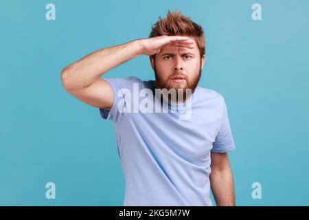 Portrait of serious concentrated bearded man looking far away at distance with hand over head, attentively searching for bright future. Indoor studio shot isolated on blue background. Stock Photo