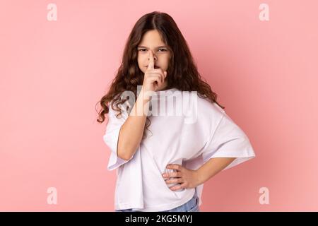 Portrait of serious concentrated little girl wearing white T-shirt holding finger on lips, making hush silence gesture, asking to keep secret. Indoor studio shot isolated on pink background. Stock Photo