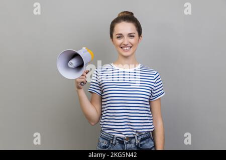 Portrait of woman wearing striped T-shirt having satisfied expression, holding megaphone in hands, looking at camera with toothy smile. Indoor studio shot isolated on gray background. Stock Photo