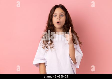Portrait of surprised little girl wearing white T-shirt raising finger up, has sudden great idea, looking at camera with open mouth. Indoor studio shot isolated on pink background. Stock Photo