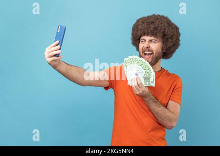 Portrait of excited crazy man with Afro hairstyle wearing orange T-shirt having video call or livestream, showing euro banknotes to subscribers. Indoor studio shot isolated on blue background. Stock Photo
