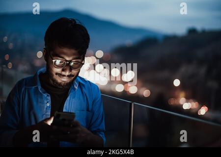 An indian man with glasses and a blue shirt using a smartphone at night on the balcony of a modern building. In the background of the night street of Stock Photo