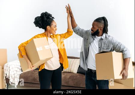 Joyful happy married african american family couple, holding boxes with things for the house, stand in living room, give five to each other, smile, rejoicing in buying their own apartment or house Stock Photo