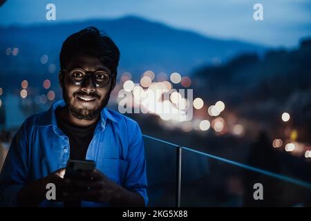 An indian man with glasses and a blue shirt using a smartphone at night on the balcony of a modern building. In the background of the night street of Stock Photo