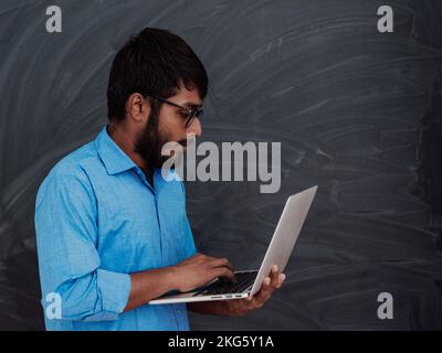 Indian smiling young student in blue shirt and glasses using laptop and posing on school blackboard background Stock Photo