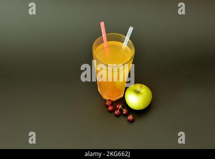 A tall glass of fruit juice with straws on a black background, next to a green apple and a handful of ripe cherries. Close-up. Stock Photo
