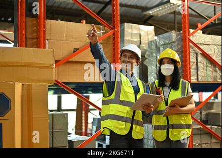 Two professional Asian warehouse workers are talking and discussing work in the warehouse, working together as a team. Factory, warehouse, industrial Stock Photo