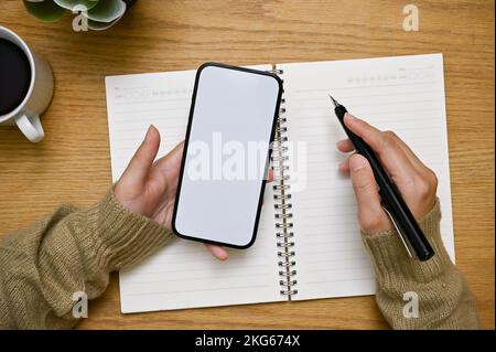 A female hands holding smartphone white screen mockup and pen, working or doing homework at her wooden desk. top view Stock Photo
