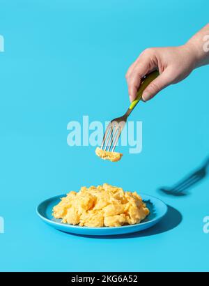 Plate with scrambled eggs in bright light minimalist on a blue table. Woman's hand taking scrambled eggs with a fork from a blue plate. Stock Photo