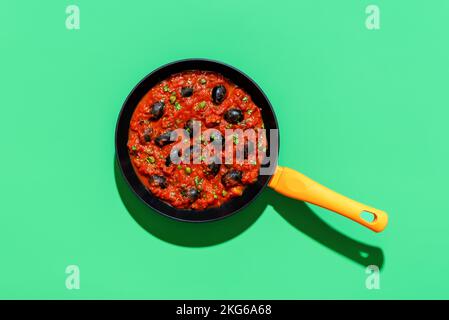 Cooking pan with pasta puttanesca sauce, minimalist on a green table. Tomato sauce, italian recipe with black olives, capers and garlic. Stock Photo