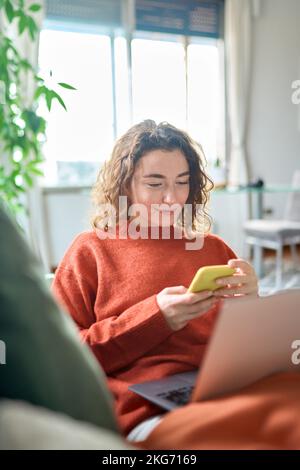 Smiling young woman relaxing on sofa using phone at cozy home, vertical. Stock Photo