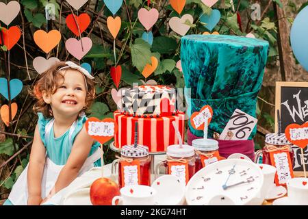 Little smiling girl 2 years old dressed as Alice in Wonderland near table with fairy tale props. Cake, Hatters hat, inscriptions Eat me and Drink me Stock Photo
