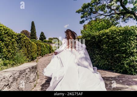a beautiful woman with long brown hair and long white dress runs along a path along beautiful bushes in the park rear view Stock Photo