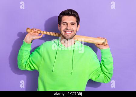 Portrait of young brunet hair man hold baseball bat professional vandal confident person wear green hoodie isolated on purple color background Stock Photo