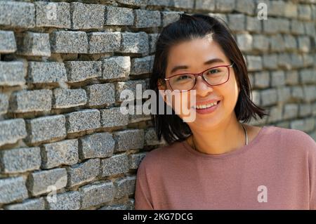 Portrait Asian middle-aged woman with smiling face, smiling, looks healthy, wears eyeglasses, stand lean on grey brick wall, perspective view, empty s Stock Photo
