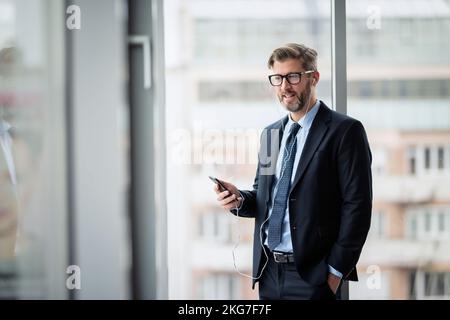 Portrait of smiling businessman using smartphone and earphones while standing at the office. Professional man wearing glasses and suit. Stock Photo