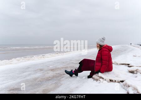 A cheerful woman in a red jacket and a warm dress sits on the shore of the winter sea. Stock Photo