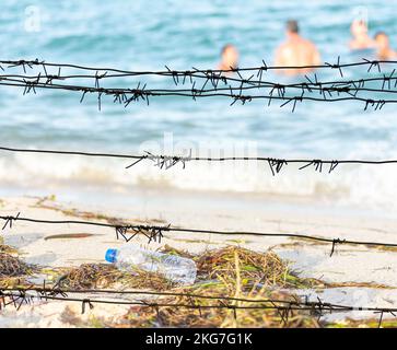 View through the barbed wire on a dirty beach filled with seaweed, garbage and swimming people Stock Photo