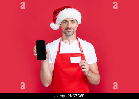 mature man in christmas santa hat and red apron hold smartphone and card. online banking Stock Photo