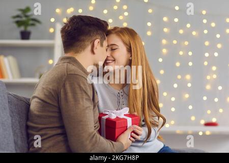 Happy couple in love sitting on couch at home, holding present and touching noses about to kiss Stock Photo