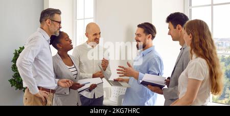 Group of business people discuss something in office standing near window with laptop and notepads. Stock Photo