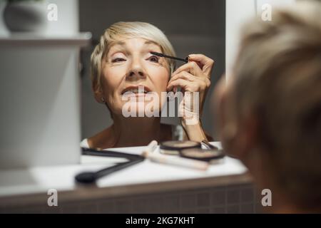 Mature woman is applying mascara in the bathroom. Stock Photo