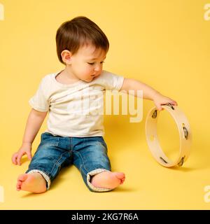 Toddler baby plays the tambourine, a child with a percussion musical instrument on a studio yellow background. Happy child musician playing hand drum. Stock Photo