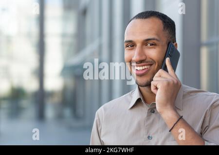 Close up photo. Handsome hispanic, african american man standing on the street and talking on the phone, waiting for an appointment, traveling. He looks at the camera, smiles Stock Photo