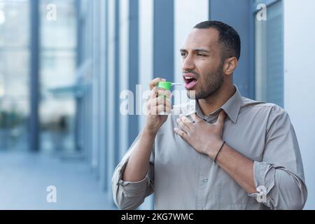 A young Latin American man clears his throat with an inhaler and medication. He stands outside, feels pain in his chest and throat, holds his throat. Stock Photo