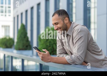 A happy handsome young African-American man is standing on the veranda of a building, holding a phone in his hands, texting with his wife, girlfriend, girlfriend, lover. Dials a message. Stock Photo