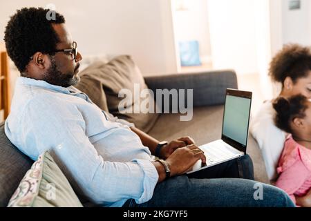 African american man using laptop while sitting on sofa with family at home Stock Photo
